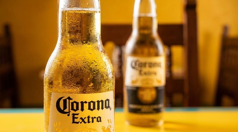Two frosty Corona Extra beer bottles, one in focus, on a yellow table with a blurred background.