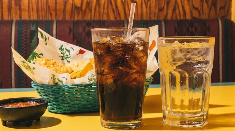 A basket of tortilla chips and salsa, with a glass of soda and water on a yellow table.