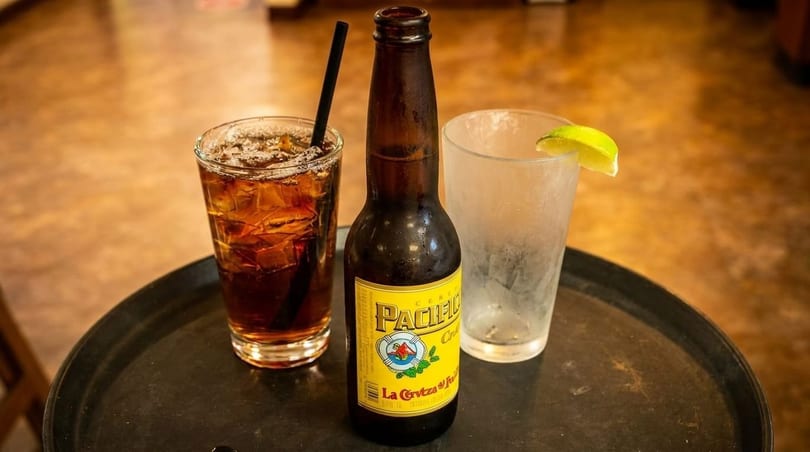 A Pacifico beer bottle, a glass of iced tea, and an empty glass with a lime wedge on a tray.