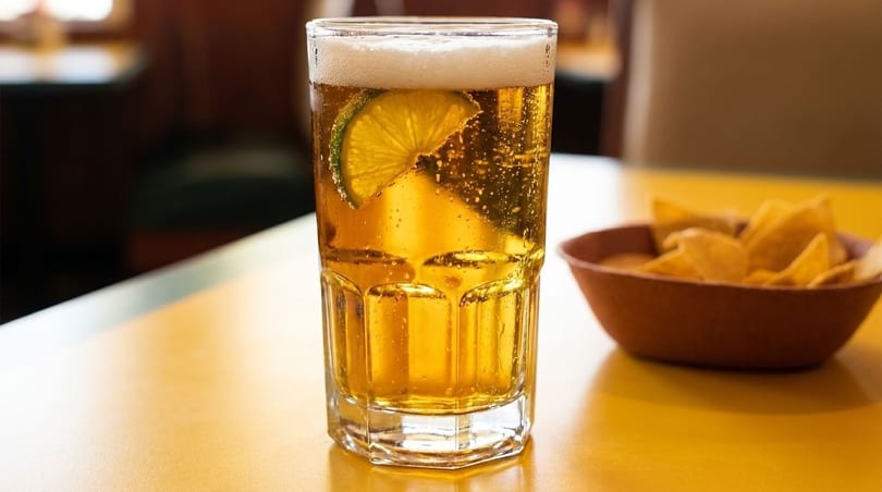 A tall glass of beer with a lime wedge and a bowl of tortilla chips on a yellow table.