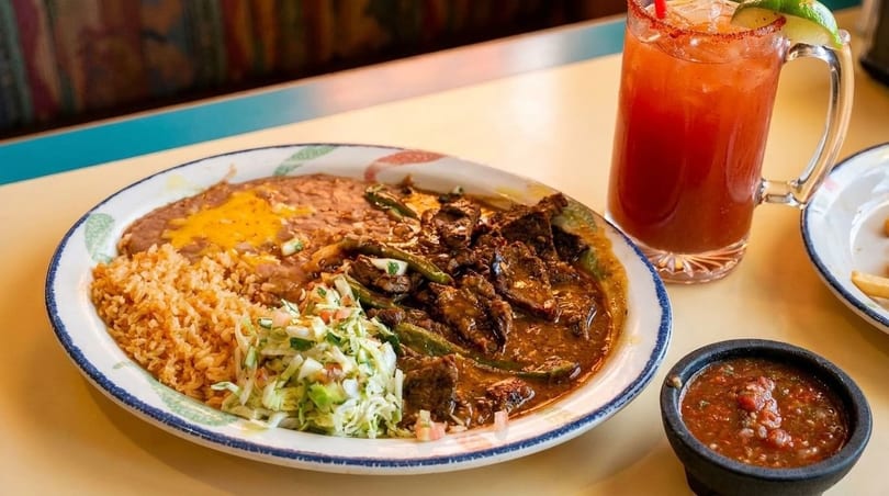 A plate of carne guisada with rice, refried beans, and coleslaw, alongside a michelada and salsa.