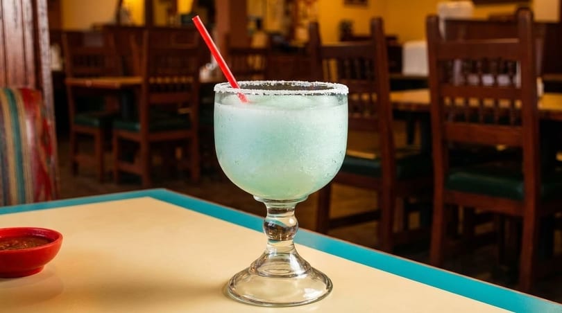 A frozen blue margarita with a red straw and salt rim sits on a table in a restaurant.