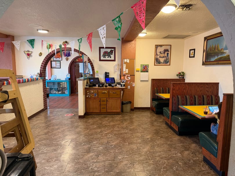 A colorful Mexican restaurant interior with festive papel picado banners, booths, and a counter area.