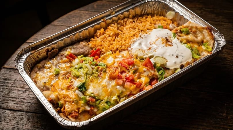 Mexican food in a foil takeout container with rice, beans, enchiladas, guacamole, and sour cream.