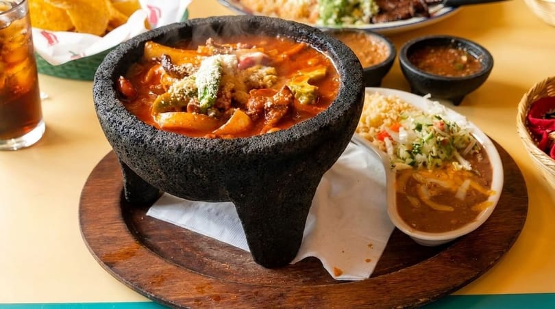 Steaming molcajete filled with a red stew, served with rice, beans, and salsa.