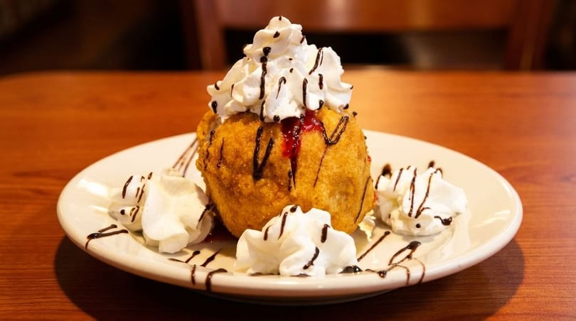 Golden-fried ice cream dessert topped with whipped cream, chocolate syrup, and raspberry sauce on a white plate.
