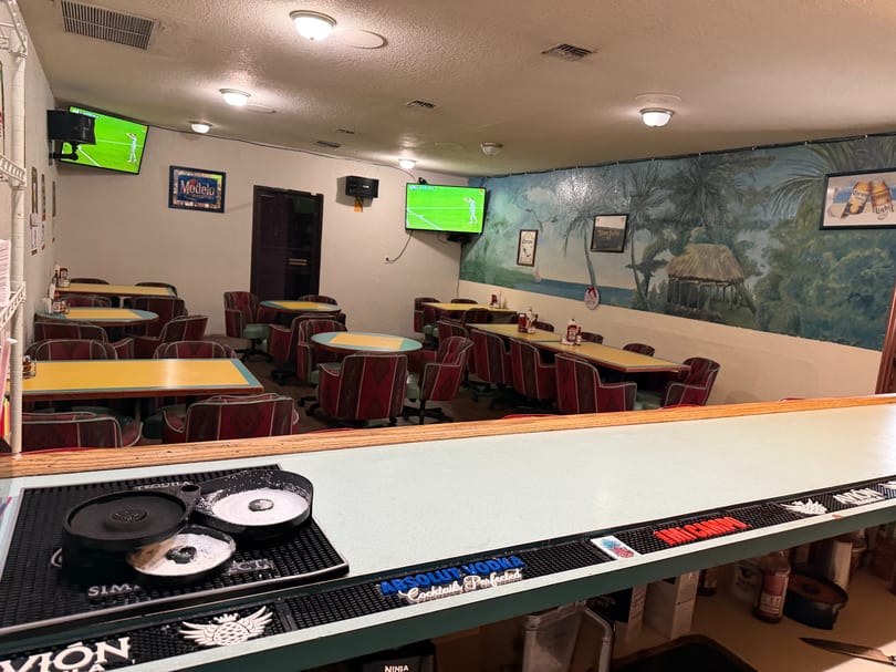 Interior of a casual restaurant with red chairs, yellow tables, a bar, and TVs showing soccer.