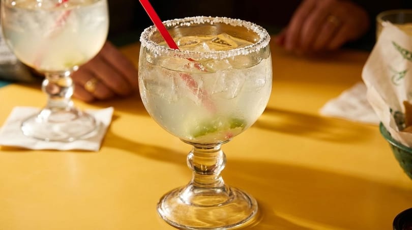 Two salt-rimmed margaritas with red straws and lime wedges on a yellow table.
