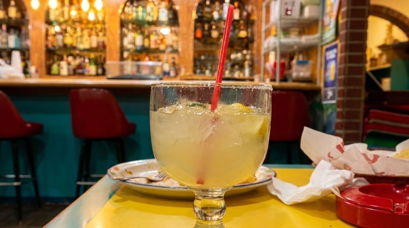 Margarita with a red straw on a yellow table, with a bar and red stools in the background.