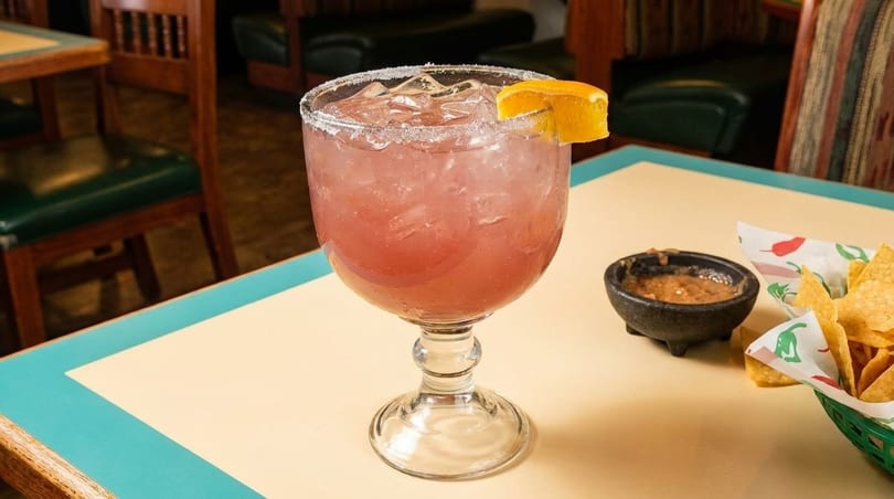 Pink margarita with an orange slice, chips, and salsa on a table in a restaurant.